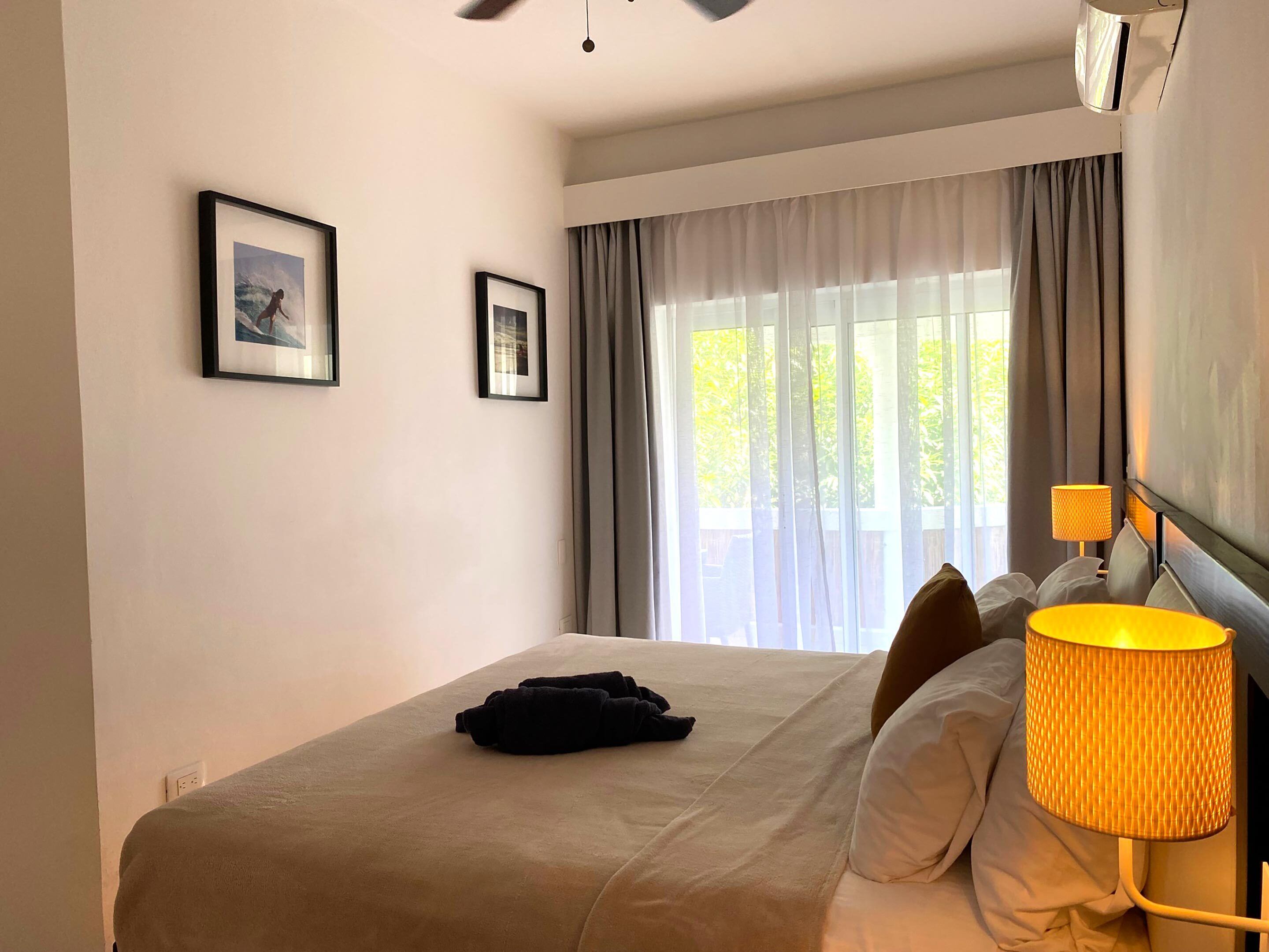 Habitación de hotel con cama, dos cuadros en la pared, una ventana con cortinas y una lámpara de noche.