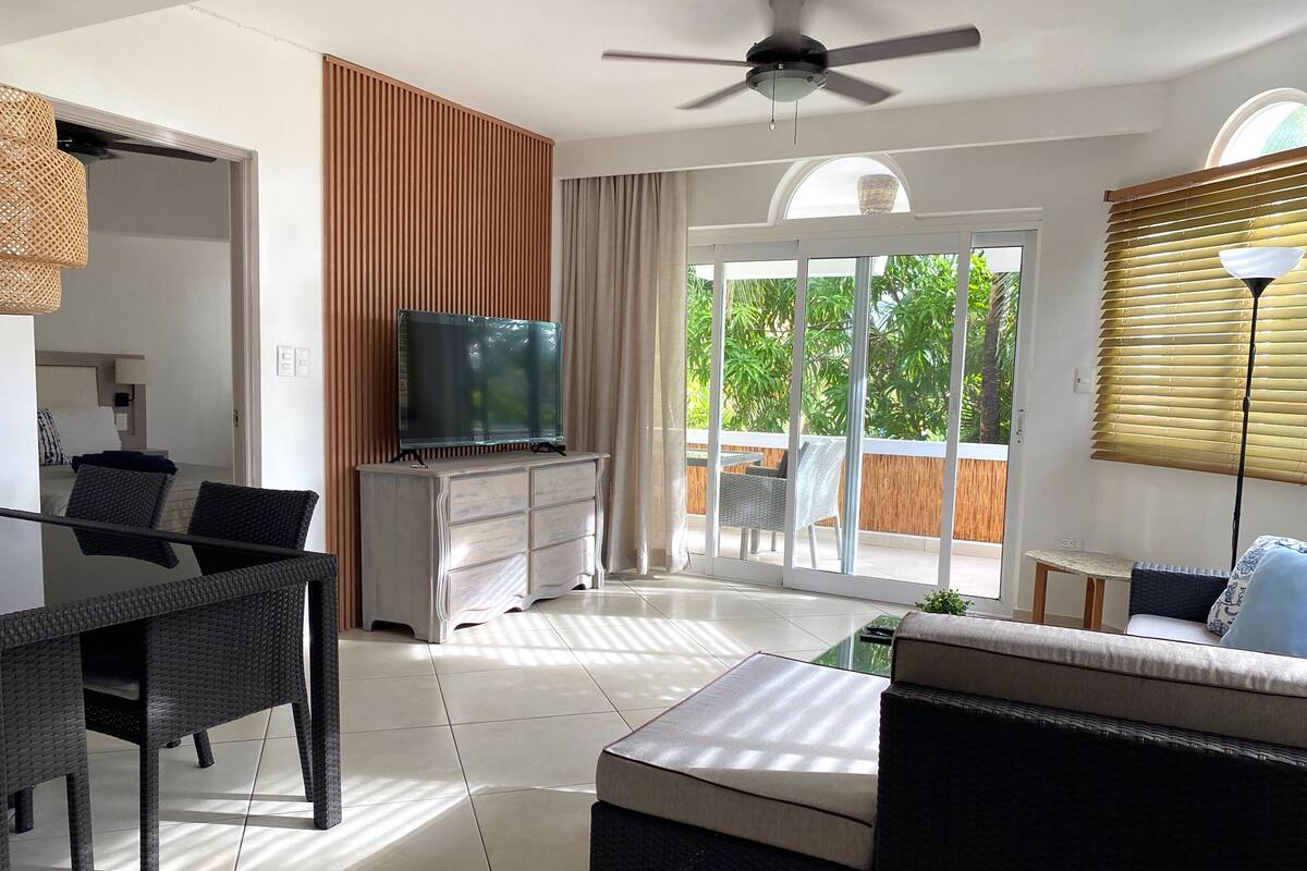 A modern living room with a television on a cabinet, a sofa, a dining table with chairs, and a balcony with a view of greenery.