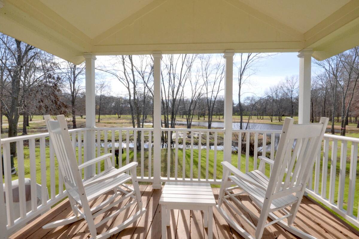 A covered porch with white wooden chairs and a bench, overlooking a grassy area with trees and a small body of water.