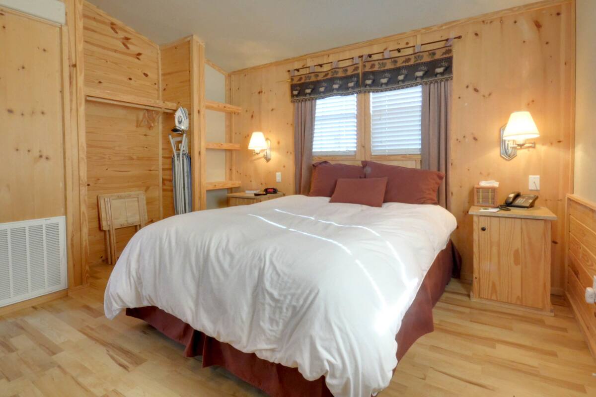 A wooden room with a bed, two bedside tables, a heater, and a window with curtains.