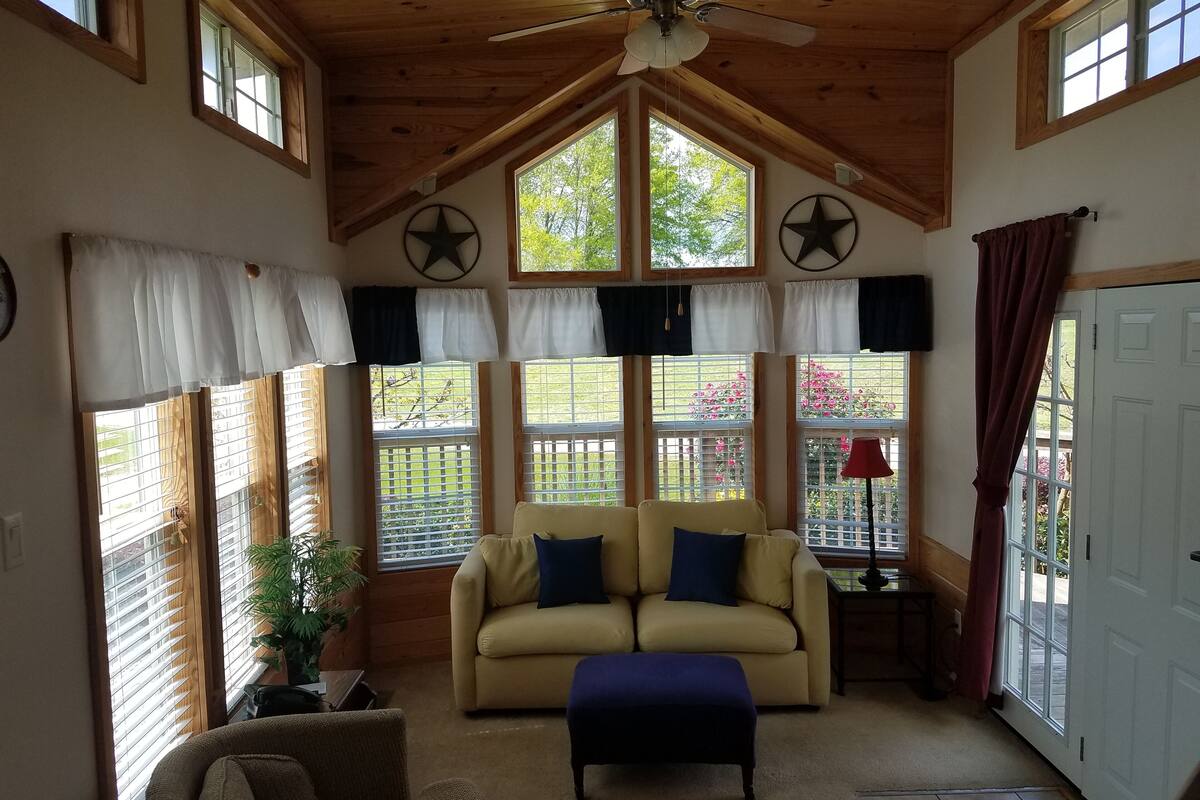 A cozy room with a wooden ceiling, a beige sofa, a blue ottoman, and a window with a view of a garden.