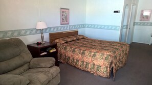 A single bed with a patterned bedspread, a nightstand with a lamp and clock, a sofa, and a framed picture on the wall. - White Valley Motel (St. Charles)