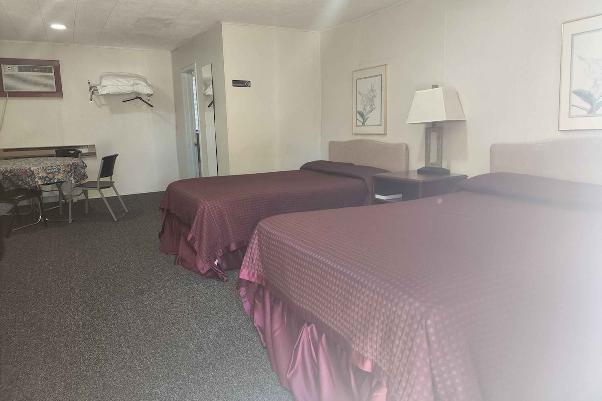Ein Hotelzimmer mit zwei Betten, einem Schreibtisch mit Stuhl, einem Tisch mit gemustertem Tischläufer und einem gerahmten Bild an der Wand.