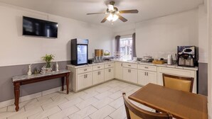 A compact kitchen with white cabinets, a marble countertop, a ceiling fan, a flat-screen TV, and a coffee machine.