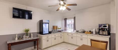 A compact kitchen with white cabinets, a marble countertop, a ceiling fan, a flat-screen TV, and a coffee machine.