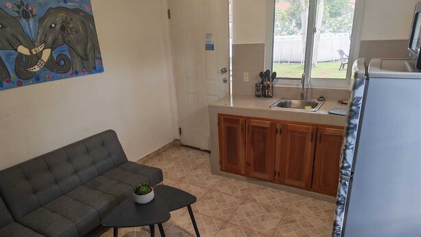 A compact living space with a sofa, a small table, a kitchen area with wooden cabinets, and a window with a view of the outdoors. - Little Italy B&B (Tijeras)