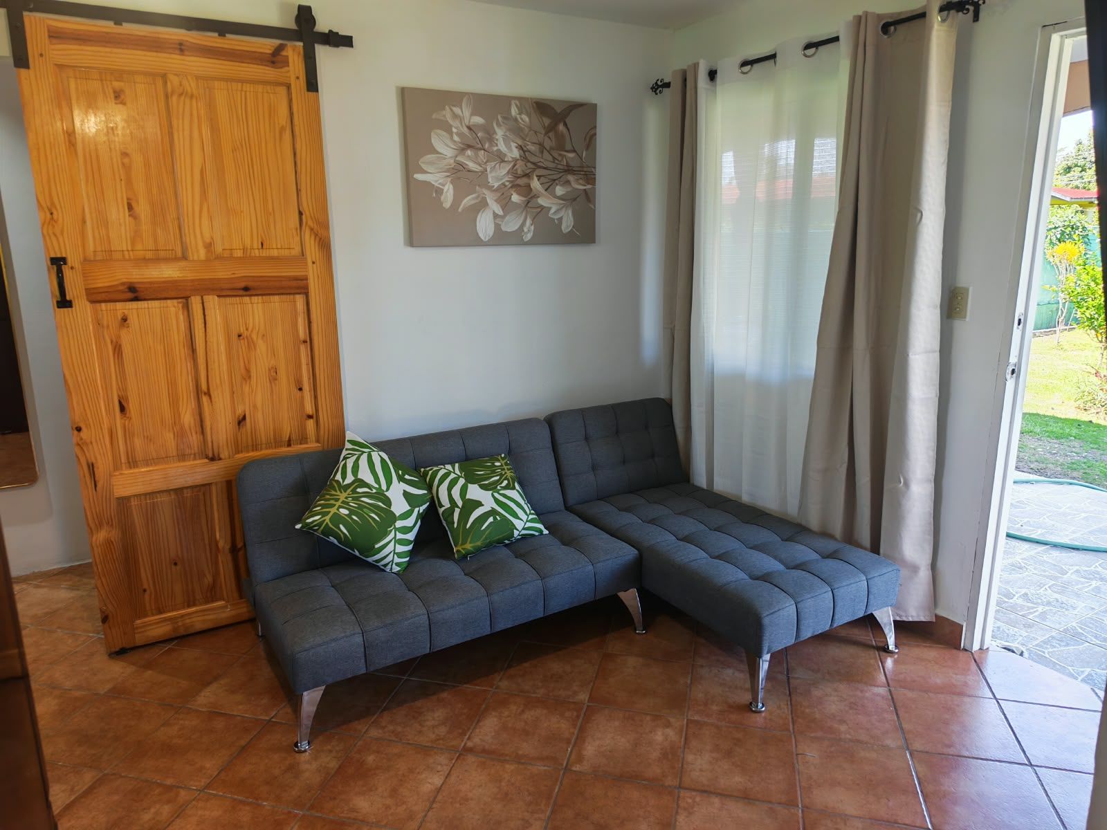 Une pièce avec un canapé gris, deux coussins à motifs verts et blancs, une porte en bois, une œuvre d’art florale accrochée au mur et une porte-fenêtre coulissante avec des rideaux.