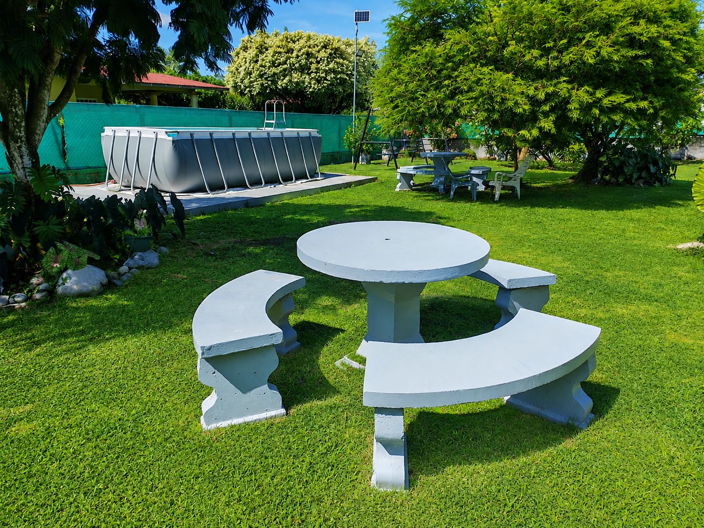 A well-maintained outdoor area with a round table and benches, surrounded by lush greenery and a swimming pool.