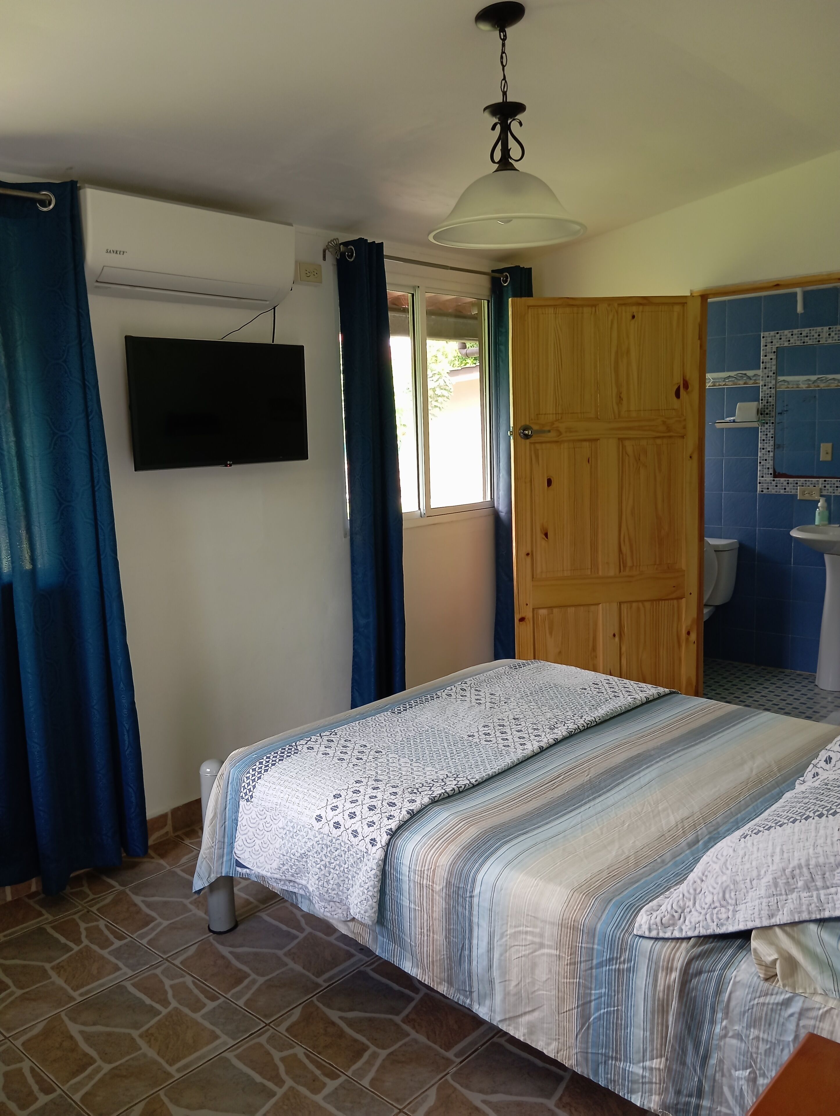 Een slaapkamer met een bed, een aan de muur bevestigde televisie, een houten deur en een badkamer met betegelde wand en wastafel.