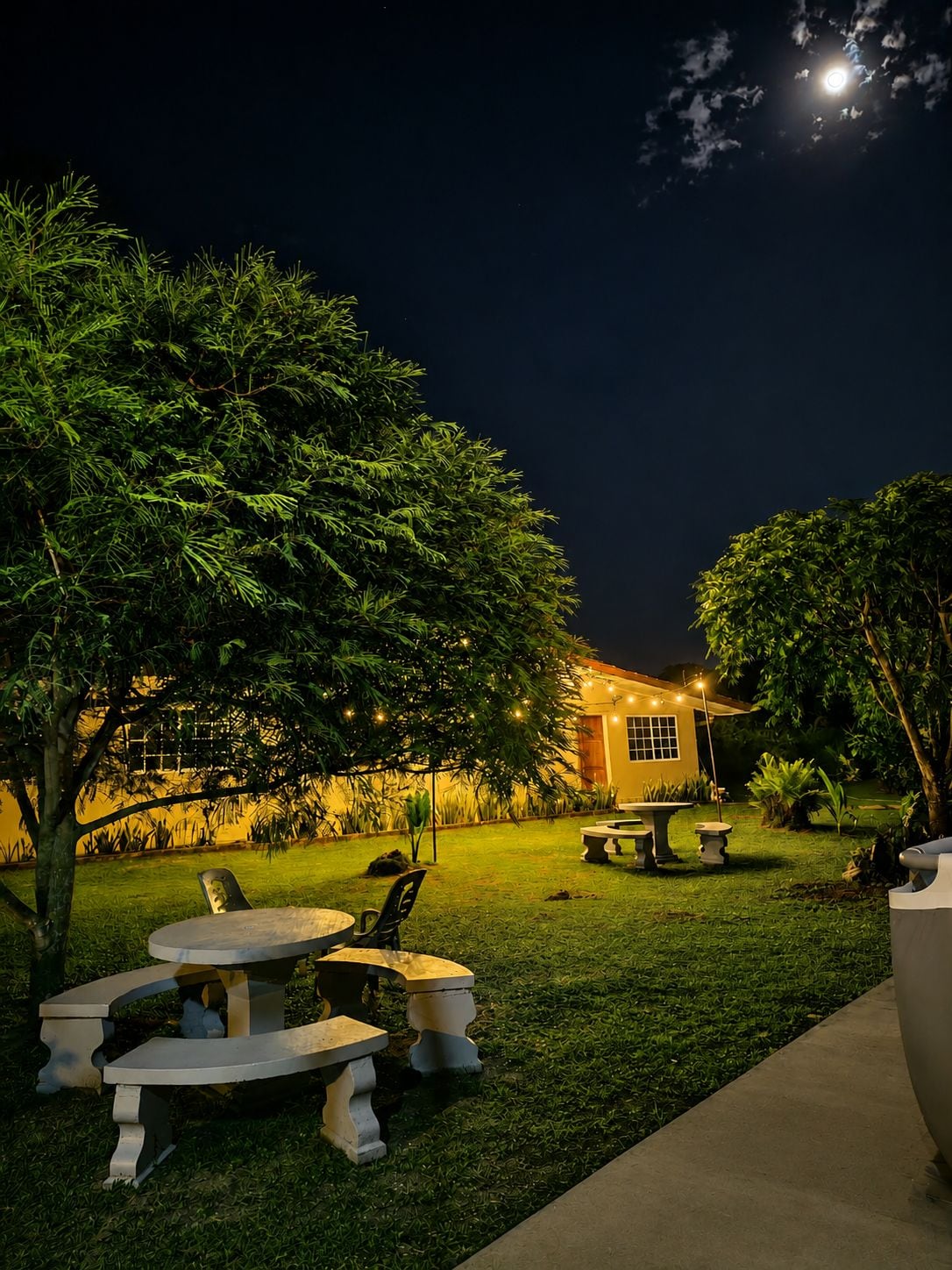 Un patio al aire libre con mesa y sillas, rodeado de follaje y un edificio al fondo.