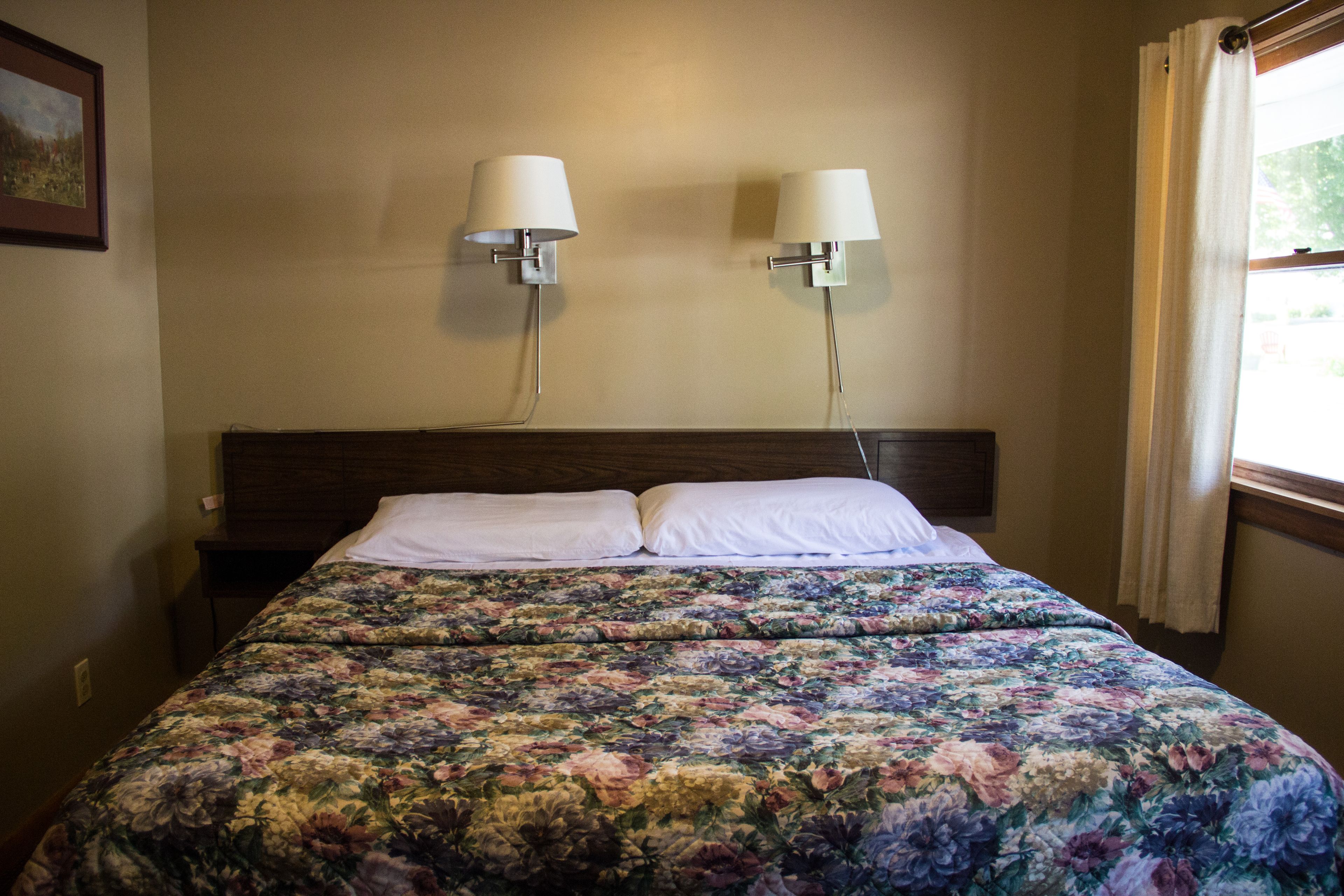 A hotel room with a floral bedspread, two wall-mounted lamps, and a window with curtains.