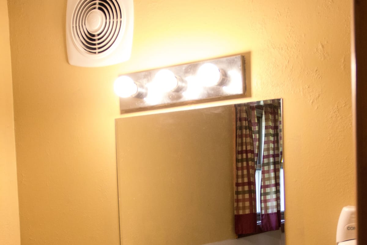 Une salle de bain équipée d’un lavabo, d’un miroir et d’un ventilateur placé au-dessus du miroir.