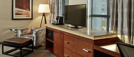 Una habitación de hotel con sofá, una mesa de centro, un televisor sobre un mueble de madera, una lámpara y una ventana con cortinas.