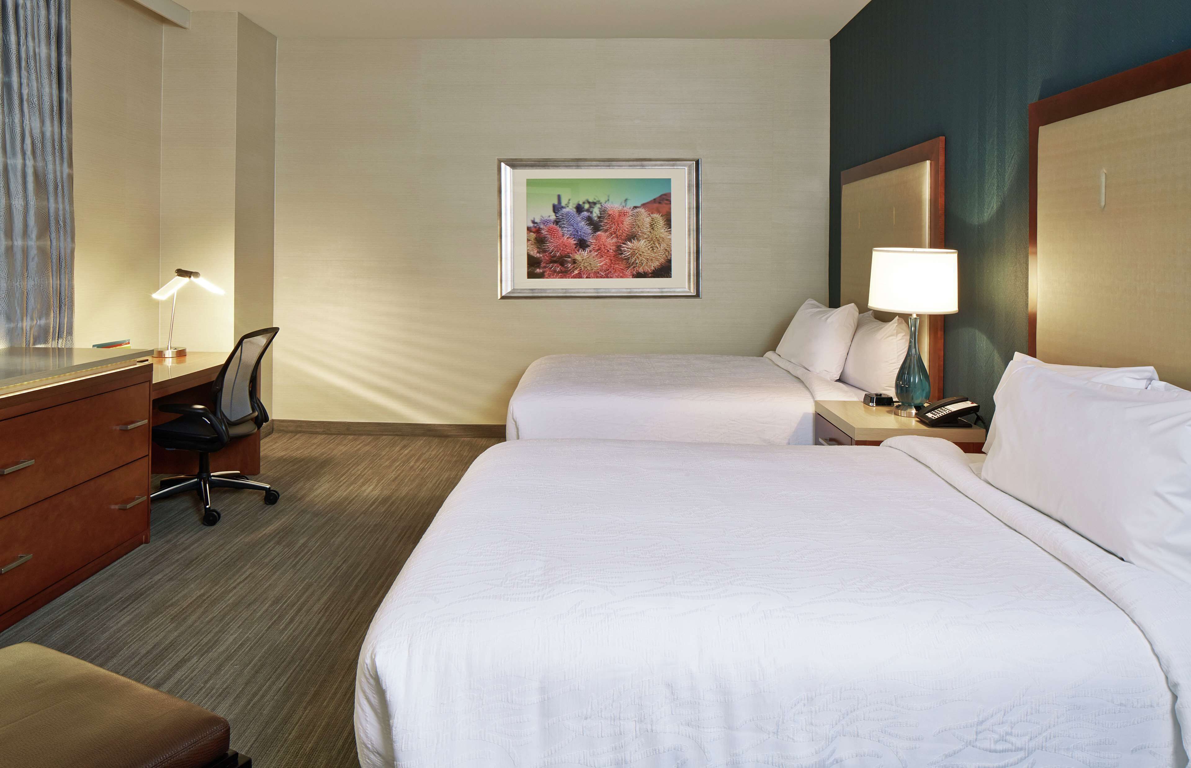A hotel room with two beds, a desk, a chair, a lamp, and a framed picture on the wall.