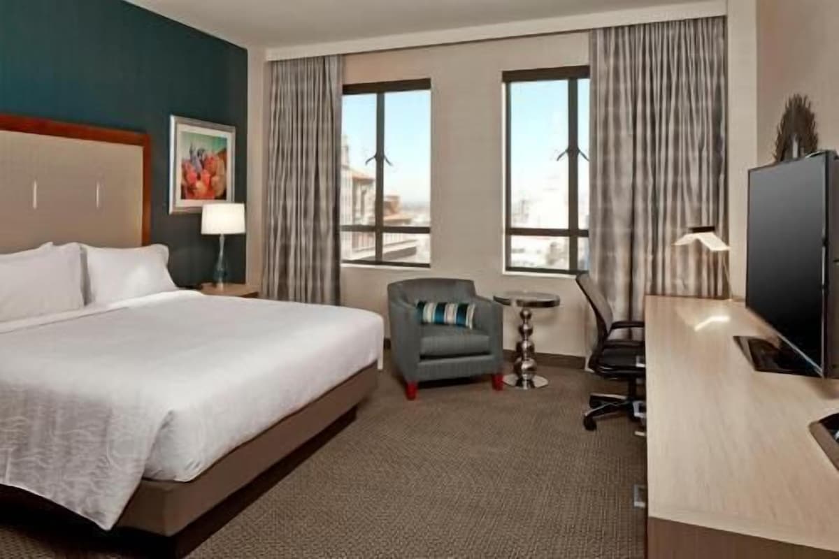 A hotel room with a large bed, a desk, a chair, and two windows with curtains.