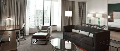 A hotel room with a large window, a sofa, a coffee table, a desk, and a bed.