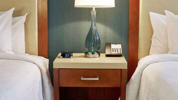 A hotel room with two beds, a bedside table with a lamp, a clock, and a phone.