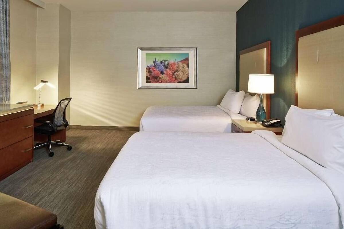 A hotel room with two beds, a desk with a chair, a lamp, a painting on the wall, and a nightstand with a lamp.