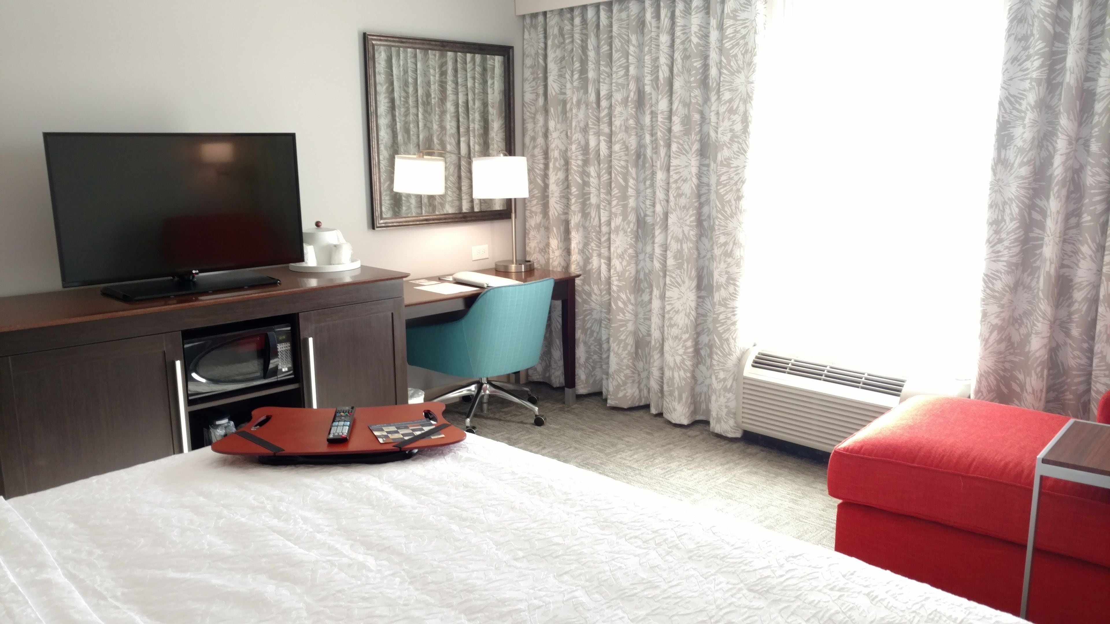 Habitación de hotel con cama, escritorio con computadora, televisor, microondas, una silla roja y ventana con cortinas.