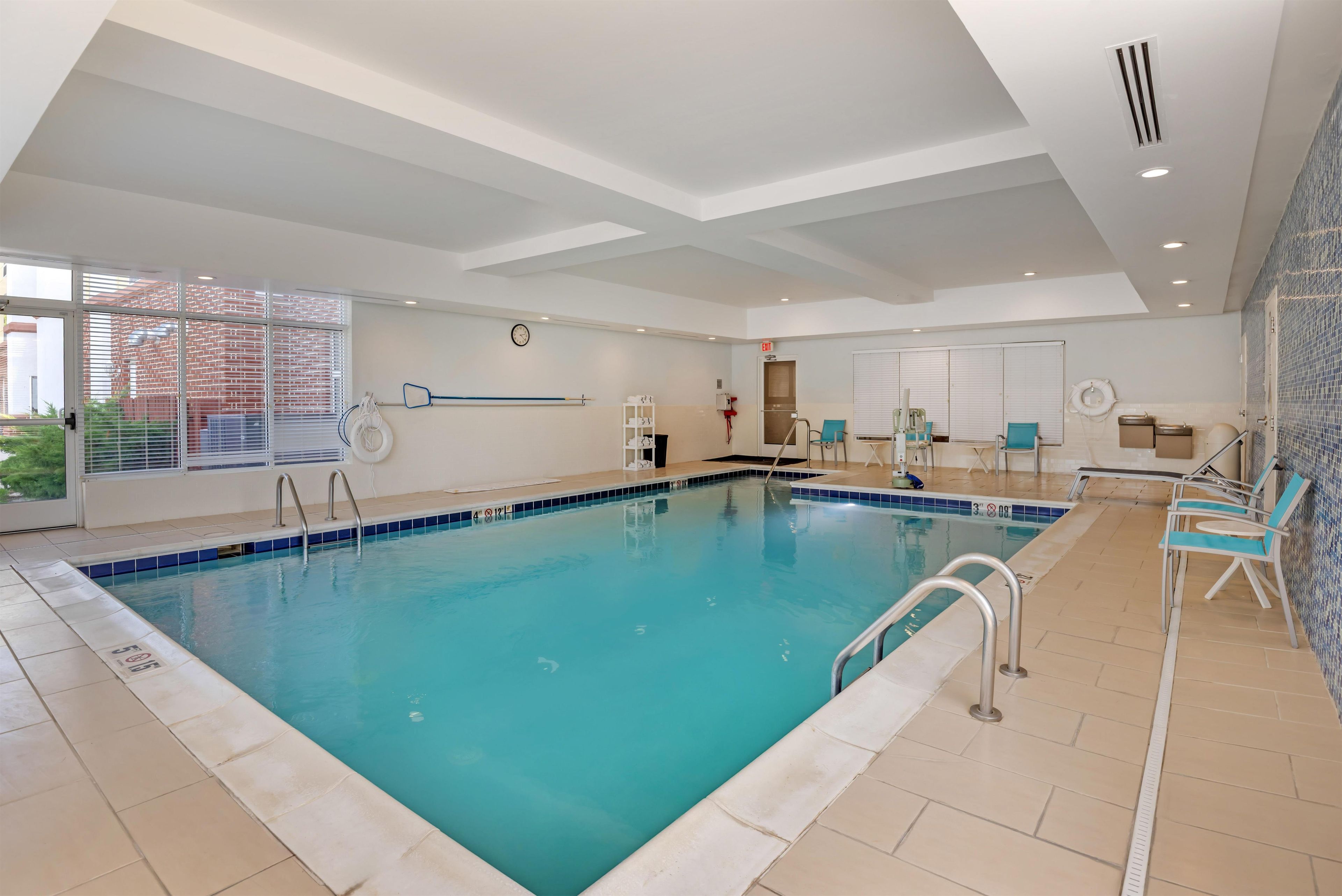 An indoor swimming pool with clear water, a clock on the wall, and a view of the outside through large windows.