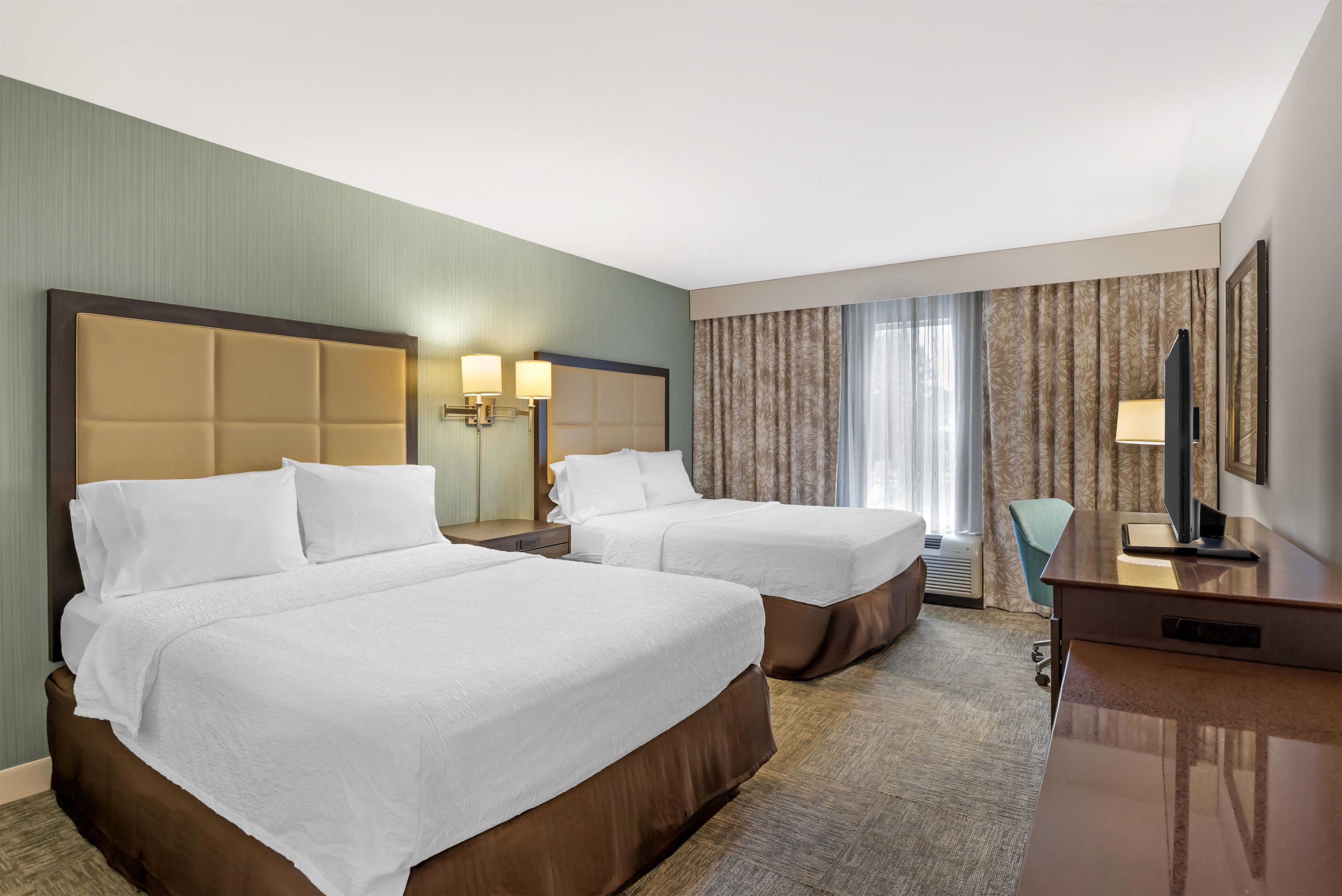 Habitación de hotel con dos camas, un escritorio, una silla, un televisor y una ventana con cortinas.