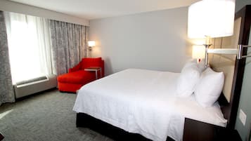 Quarto de hotel com uma cama grande, uma poltrona vermelha, uma mesa de cabeceira, um abajur e uma janela com cortinas.