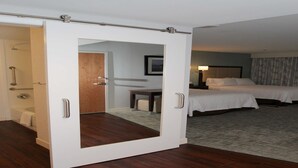 A hotel room with a wooden floor, a bed, a desk, a chair, a bathroom with a shower, and a mirror. - Hampton Inn Suffolk (Suffolk)