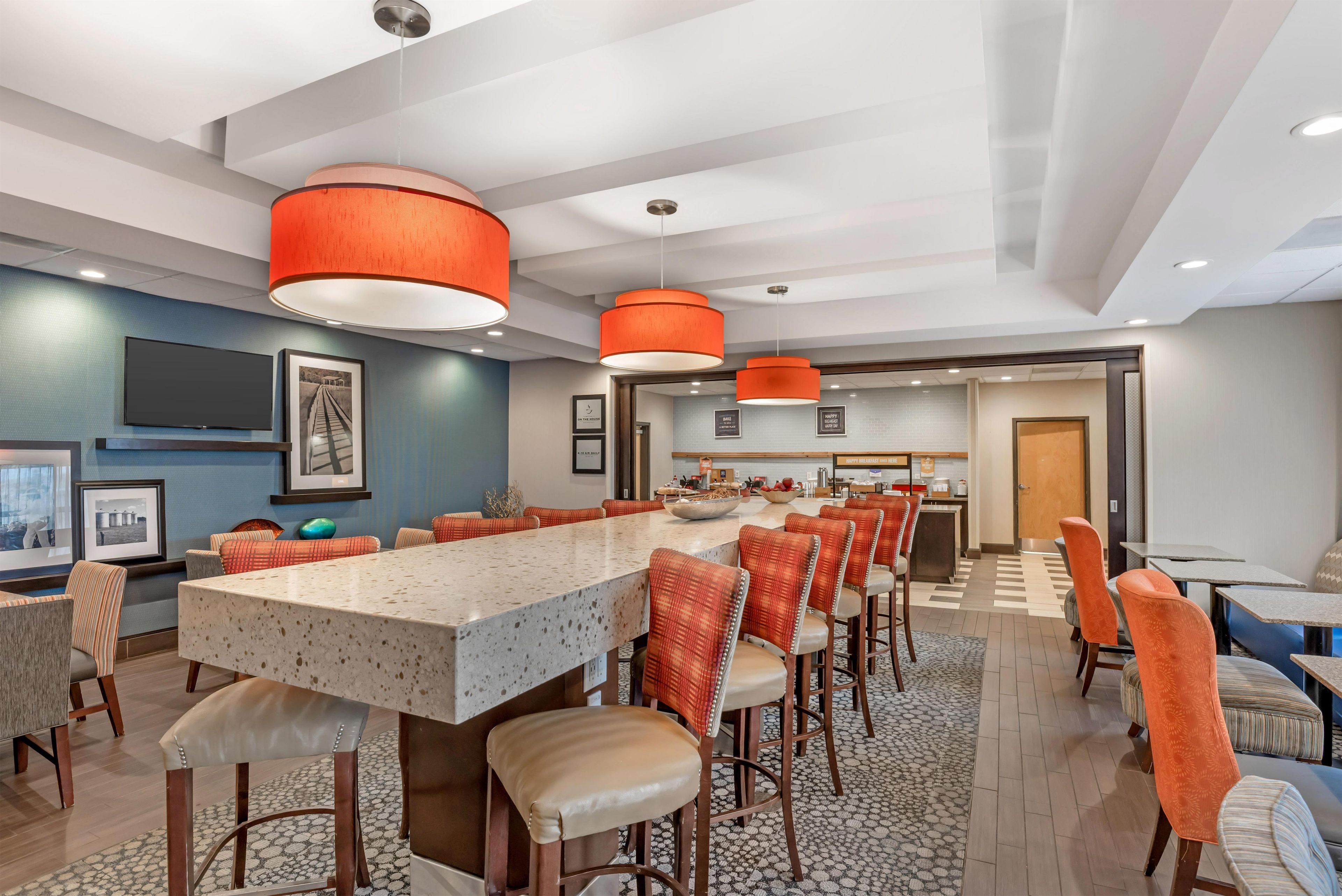 A modern hotel dining area with a long communal table, bar stools, and a kitchen in the background.