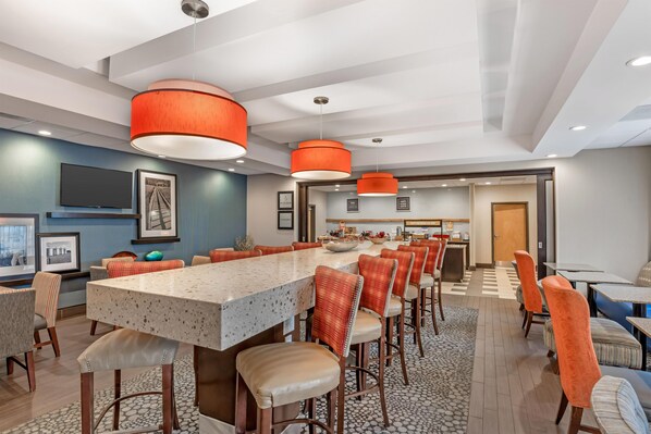 Comedor moderno de hotel con una larga mesa común, taburetes de bar y una cocina al fondo.