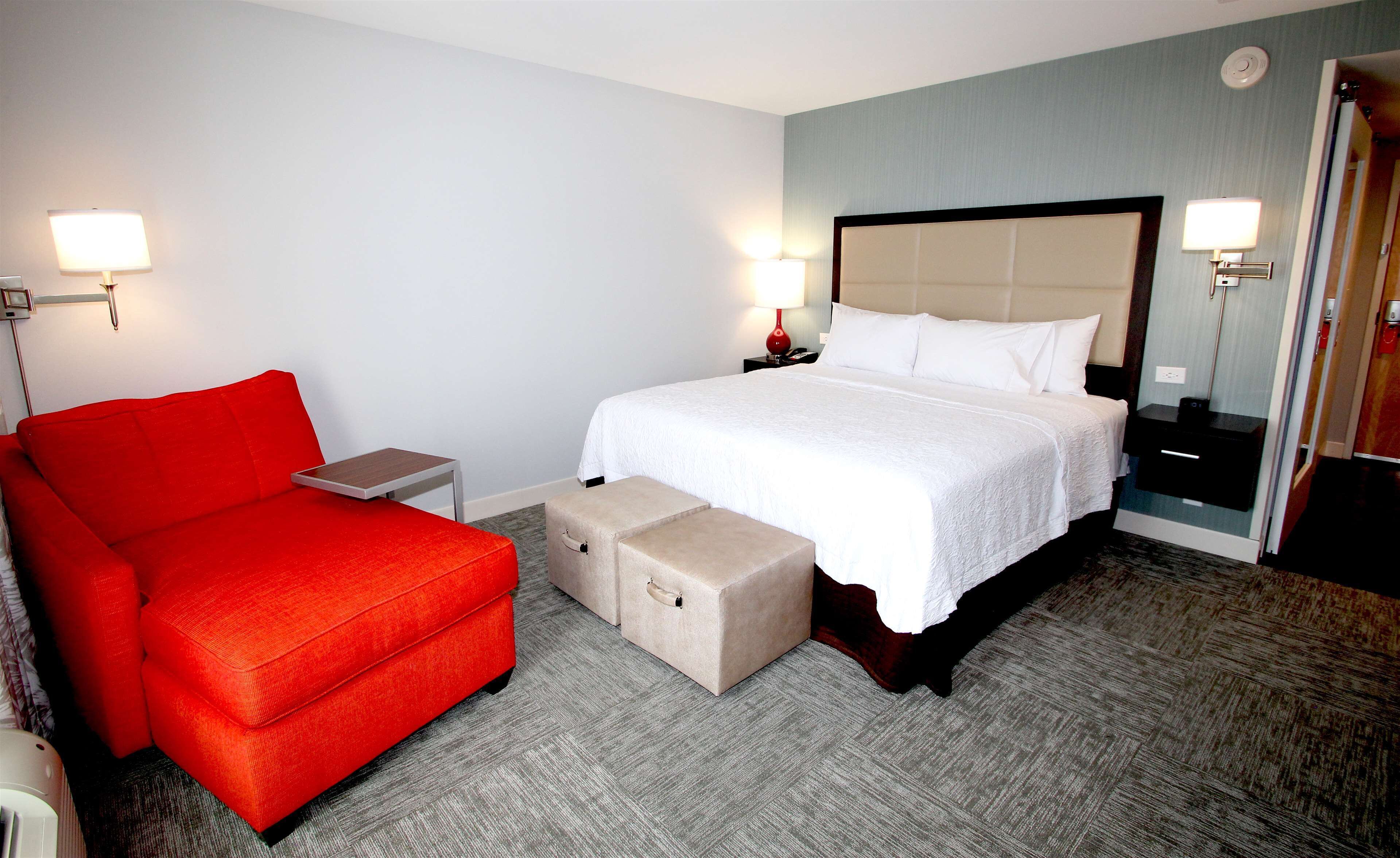 A hotel room with a bed, a red sofa, a small table, and a storage ottoman.
