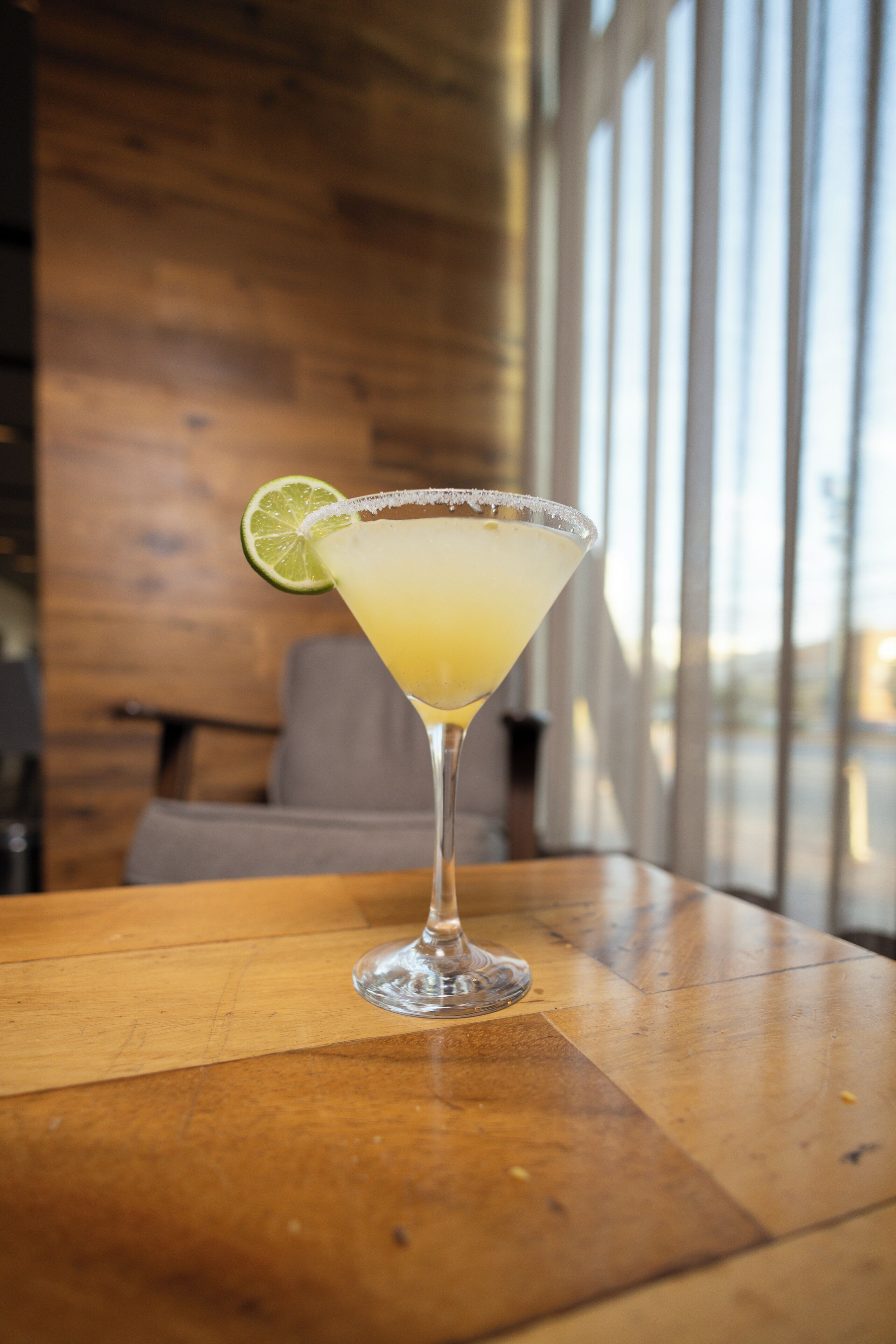 Una copa de martini con un adorno de lima sobre una mesa de madera en una habitación con ventanas amplias.