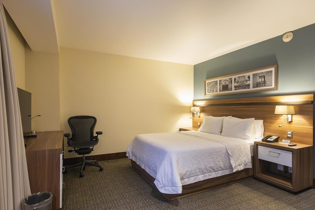 Una habitación de hotel con cama, mesita de noche, lámpara, silla y escritorio con un cubo de basura.