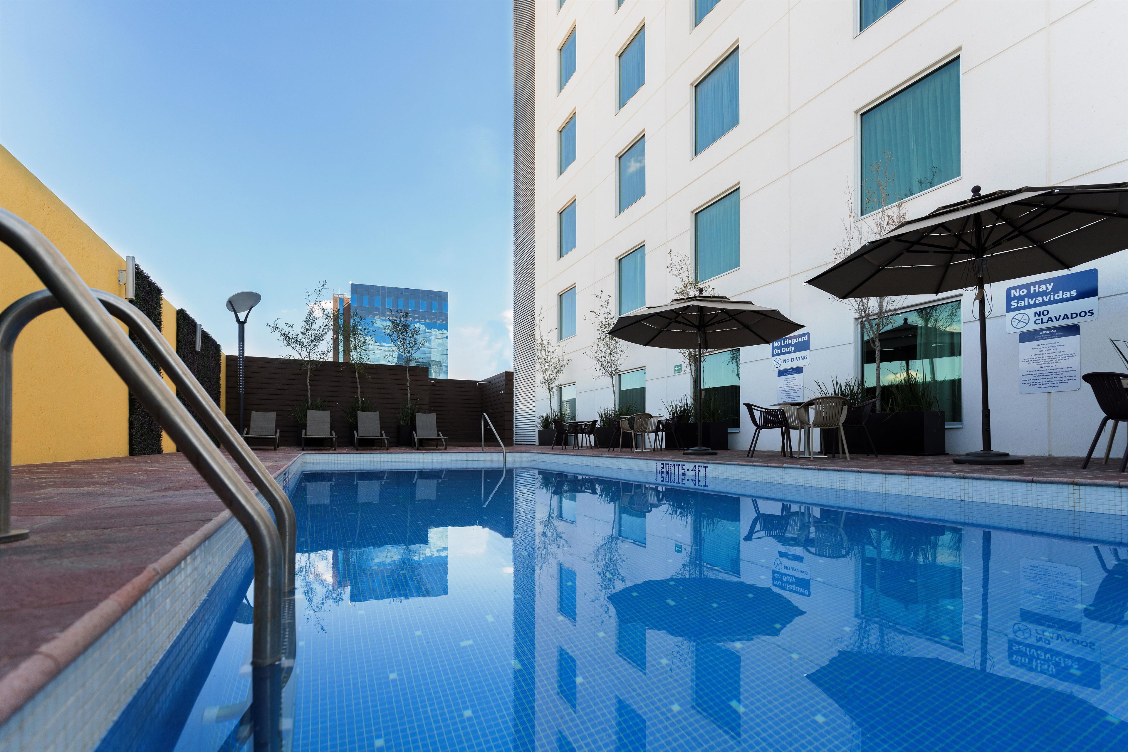 Una piscina con sillas de descanso y sombrillas, rodeada de edificios.