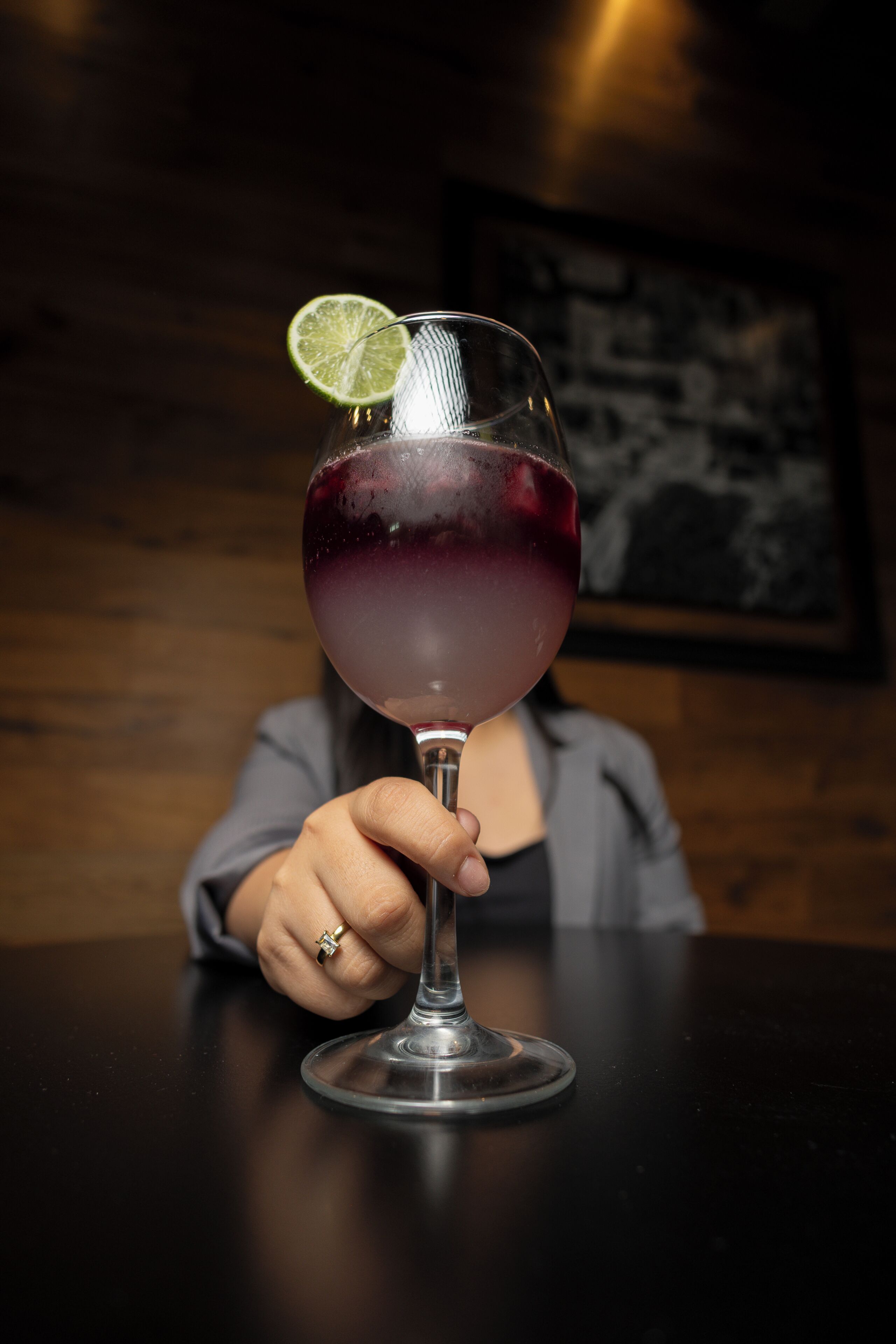Una persona sosteniendo un vaso con una rodaja de lima, sentada en una mesa con un fondo de madera oscura y un cuadro colgado en la pared.
