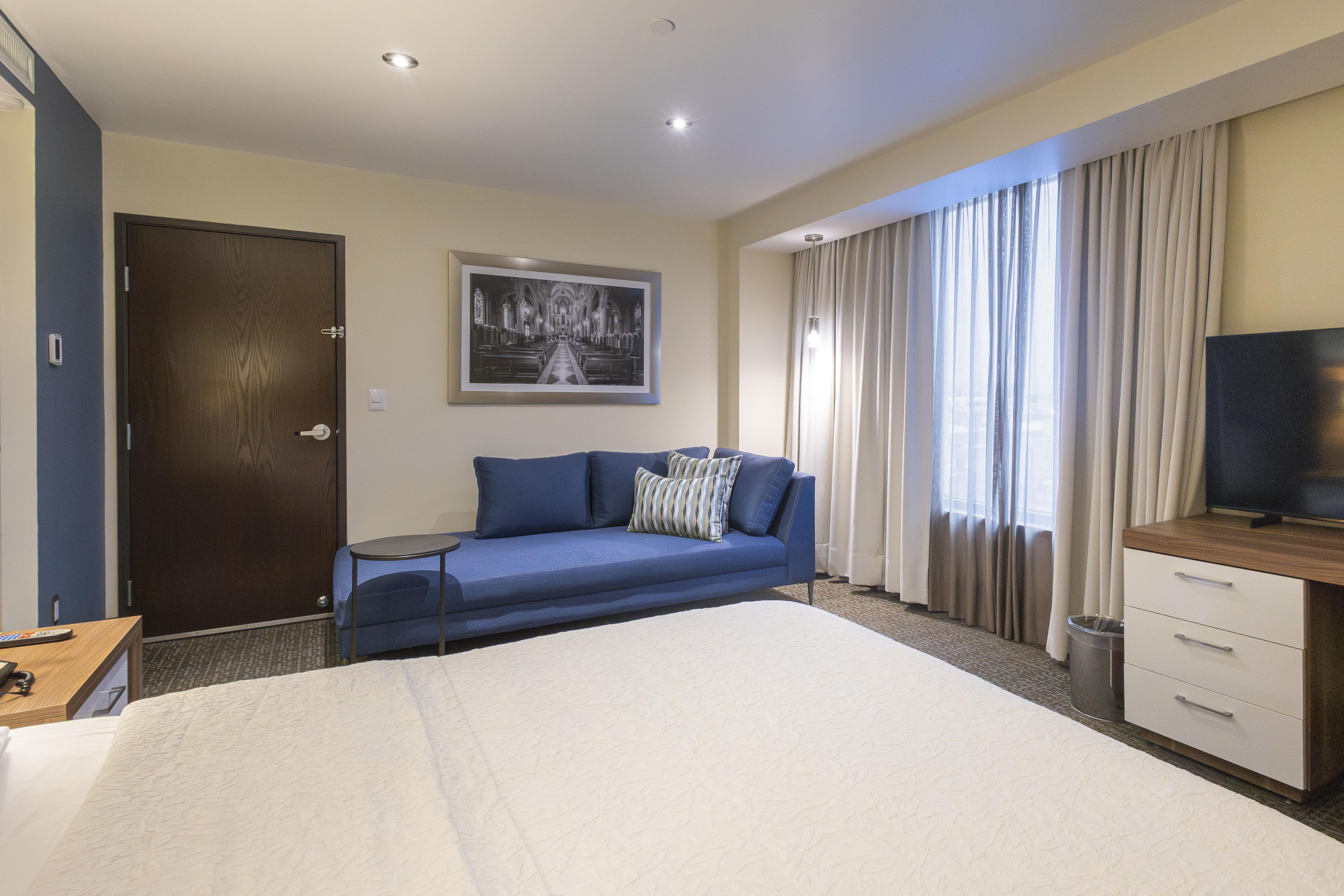Une chambre d’hôtel comprenant un lit, un canapé, un bureau avec une télévision, une porte et une fenêtre avec des rideaux.