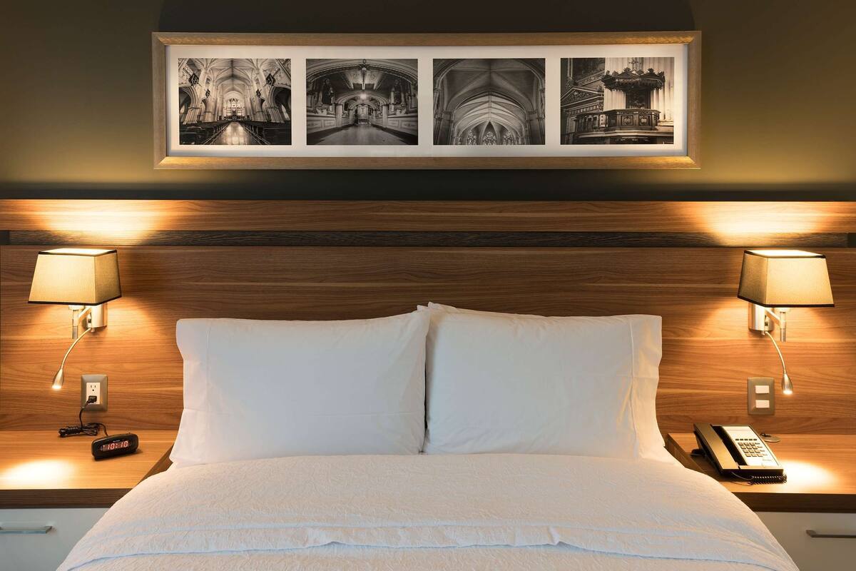 Habitación de hotel con cabecera de madera, cama con ropa de cama blanca, dos lámparas de noche, un reloj digital y un teléfono en la mesita de noche.