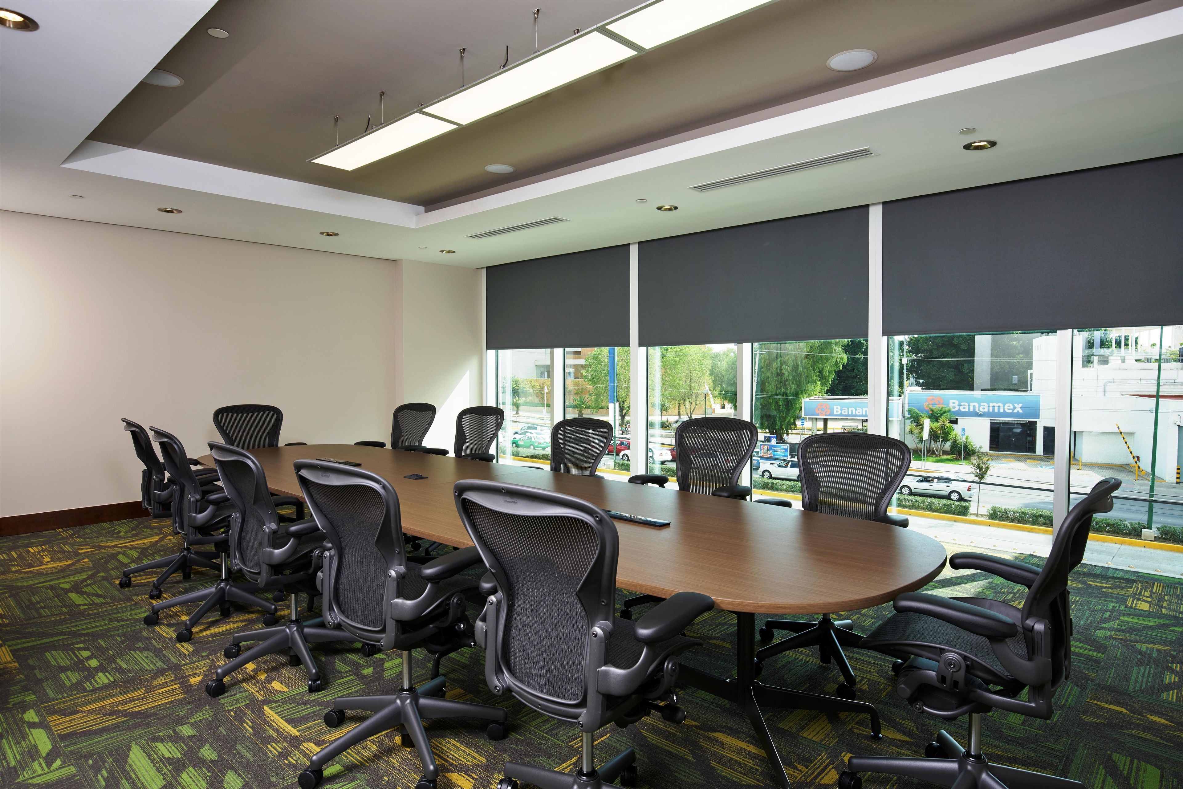 Un salón de conferencias con una gran mesa, varias sillas y amplias ventanas con persianas.