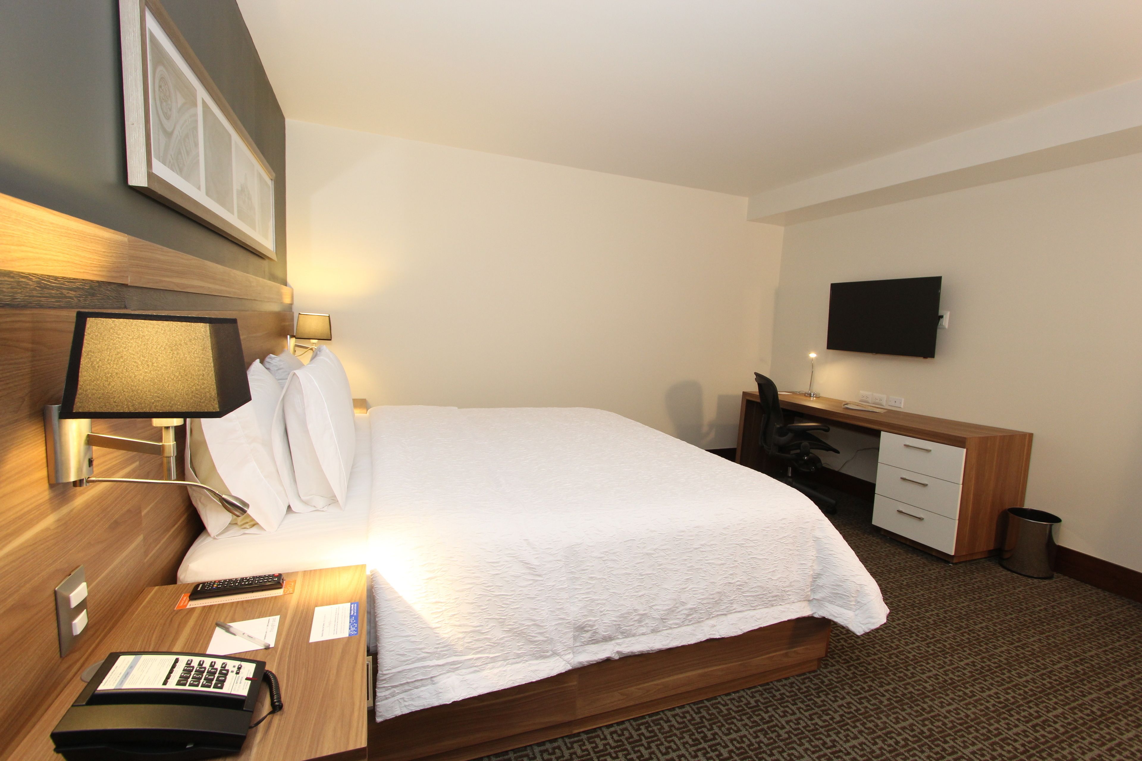 Habitación de hotel con una cama grande, un escritorio con computadora, un televisor en la pared y una lámpara en la mesita de noche.
