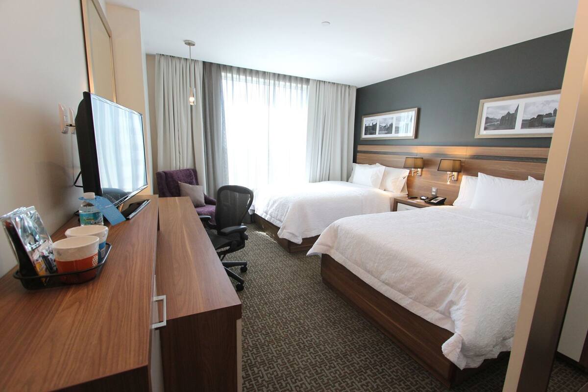 Habitación de hotel con dos camas, un escritorio con ordenador, una silla, una lámpara y una ventana con cortinas.