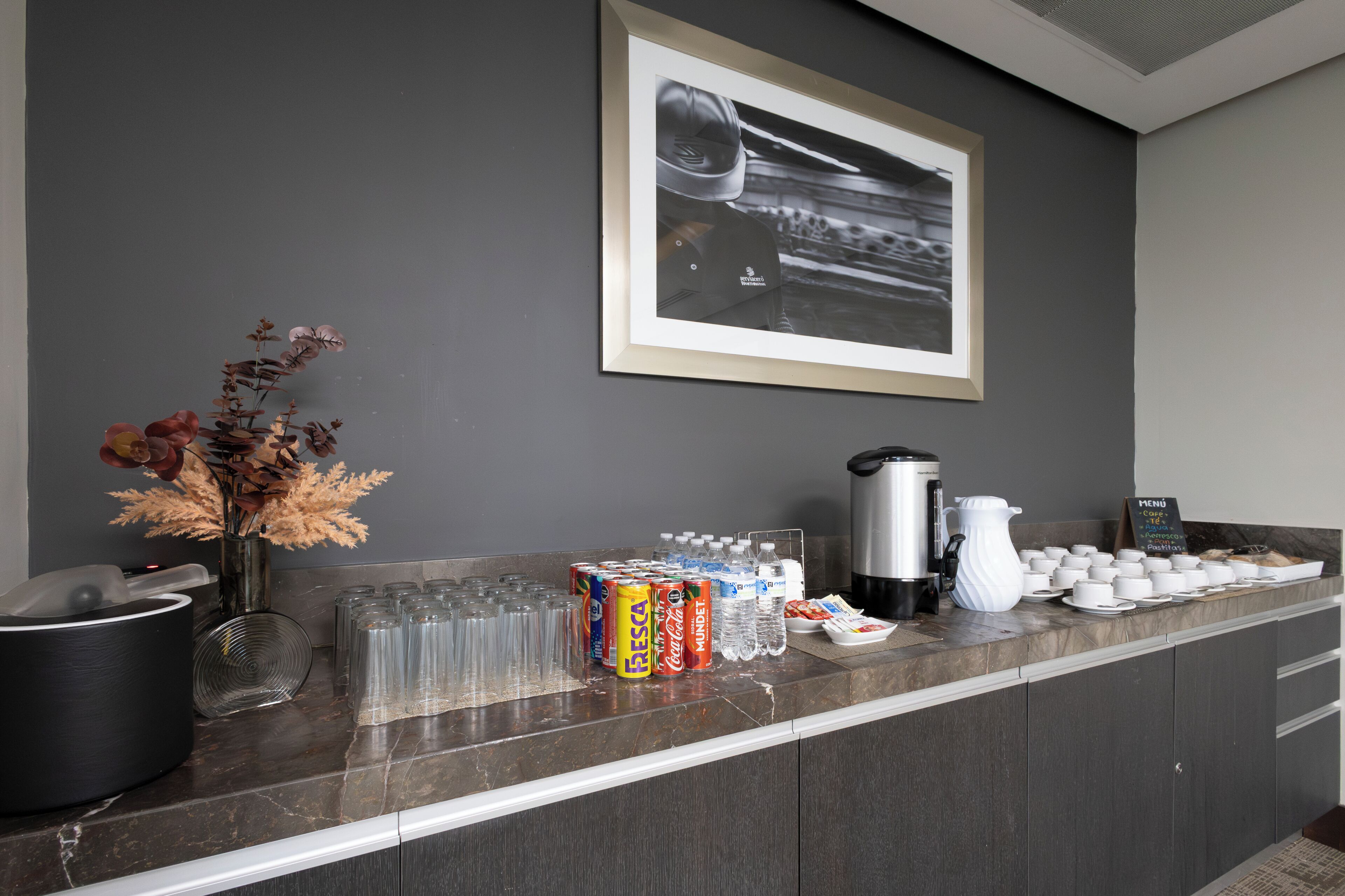 A hotel reception area with a coffee station, beverage dispensers, and a framed photograph on the wall.