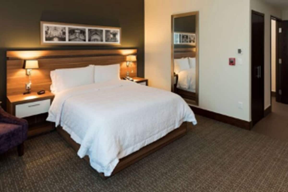Habitación de hotel con cama, lámparas de noche, mesita de noche, una silla, un espejo grande y una puerta.