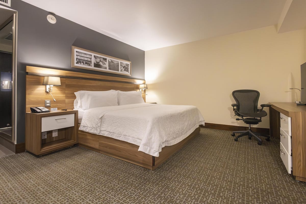 Una habitación de hotel con cama, mesita de noche, lámpara, silla, escritorio y un cuadro en la pared.