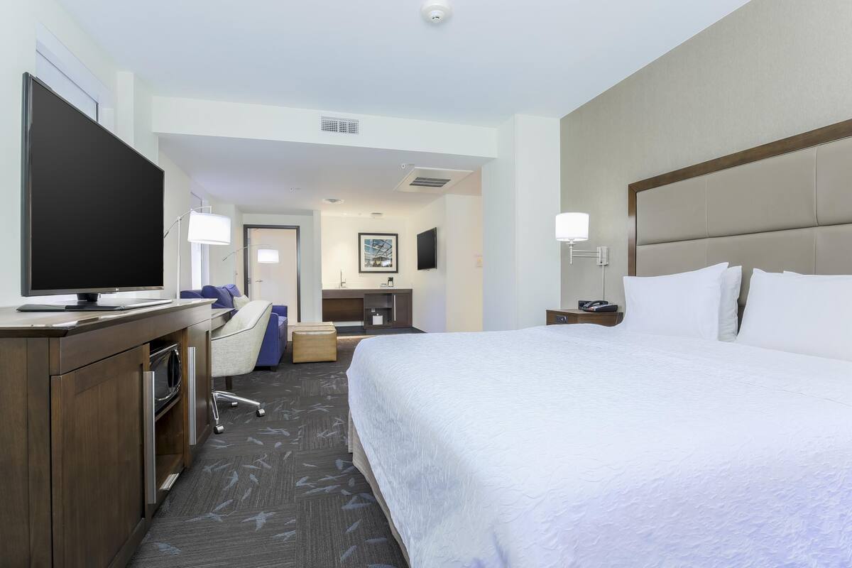 A hotel room with a bed, a desk with a TV, a chair, a desk lamp, and a nightstand.