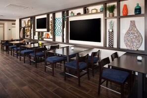 Un'area pranzo moderna con diversi tavoli e sedie, un grande televisore piatto e mensole decorative a parete.
