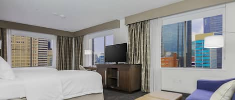 A hotel room with a bed, a TV, a sofa, and a view of city buildings.