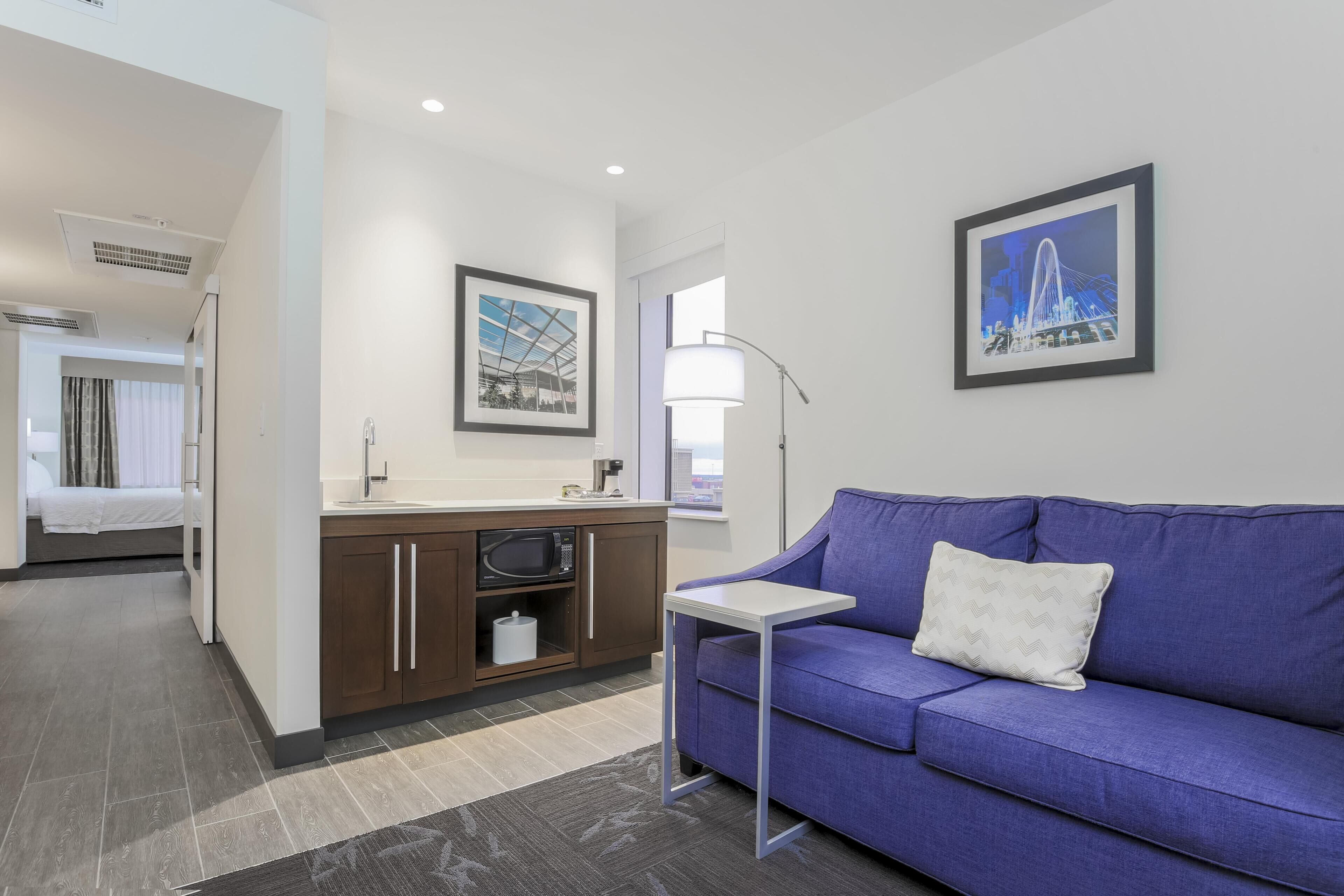 A modern hotel room with a blue sofa, a small table, a microwave, a sink, and framed pictures on the wall.