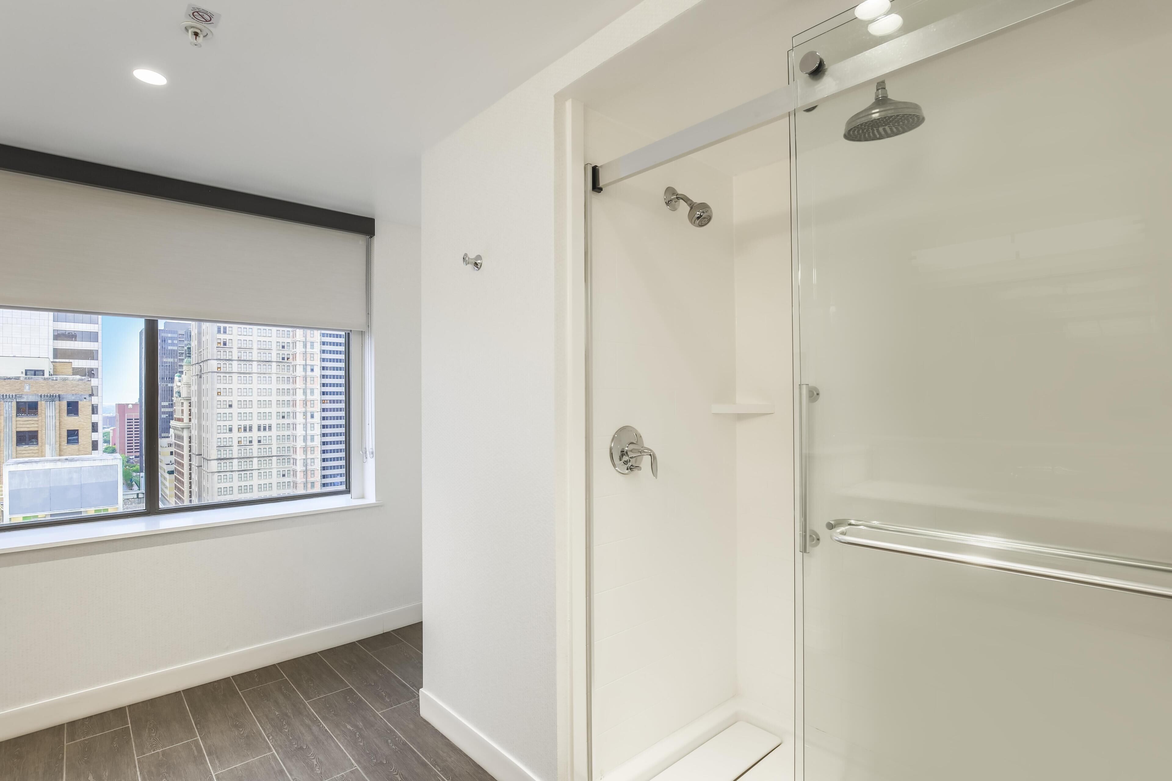 A modern bathroom with a glass shower enclosure, a window with a city view, and a dark wood floor.