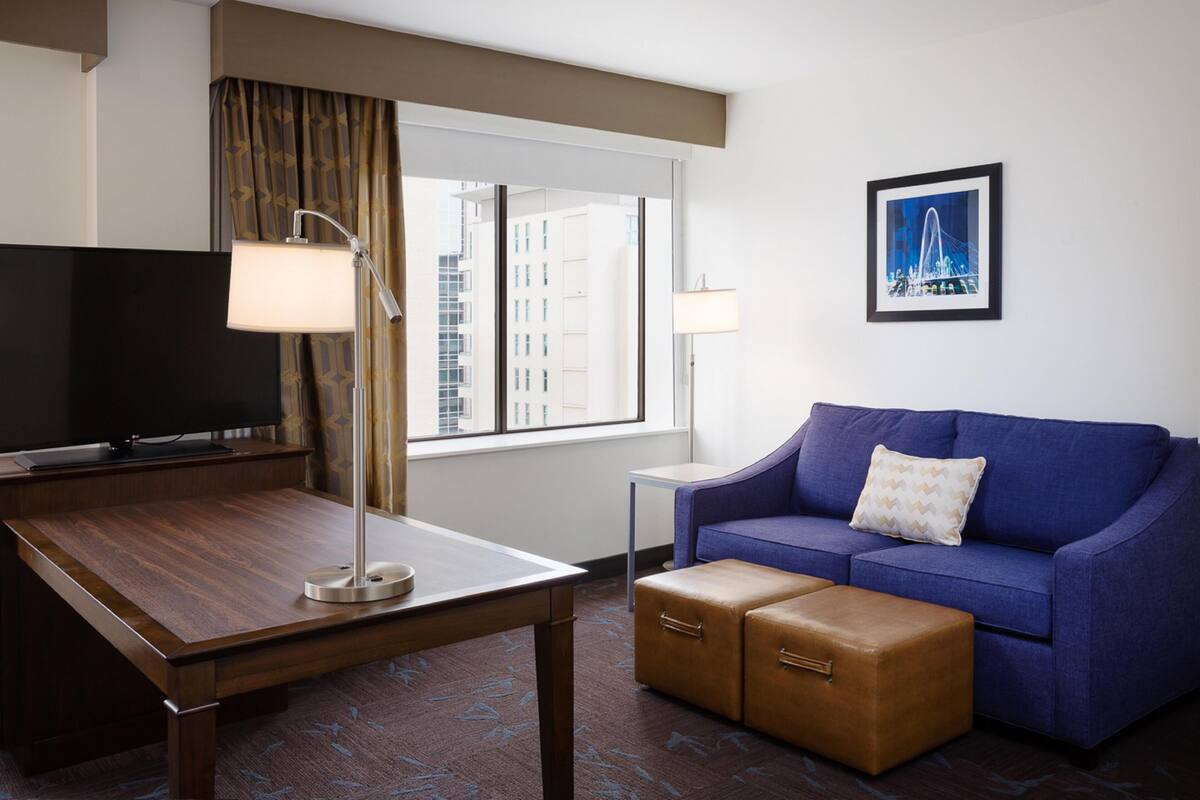 A hotel room with a wooden desk, a television, a blue sofa with a cushion, a small table, and a window with curtains.