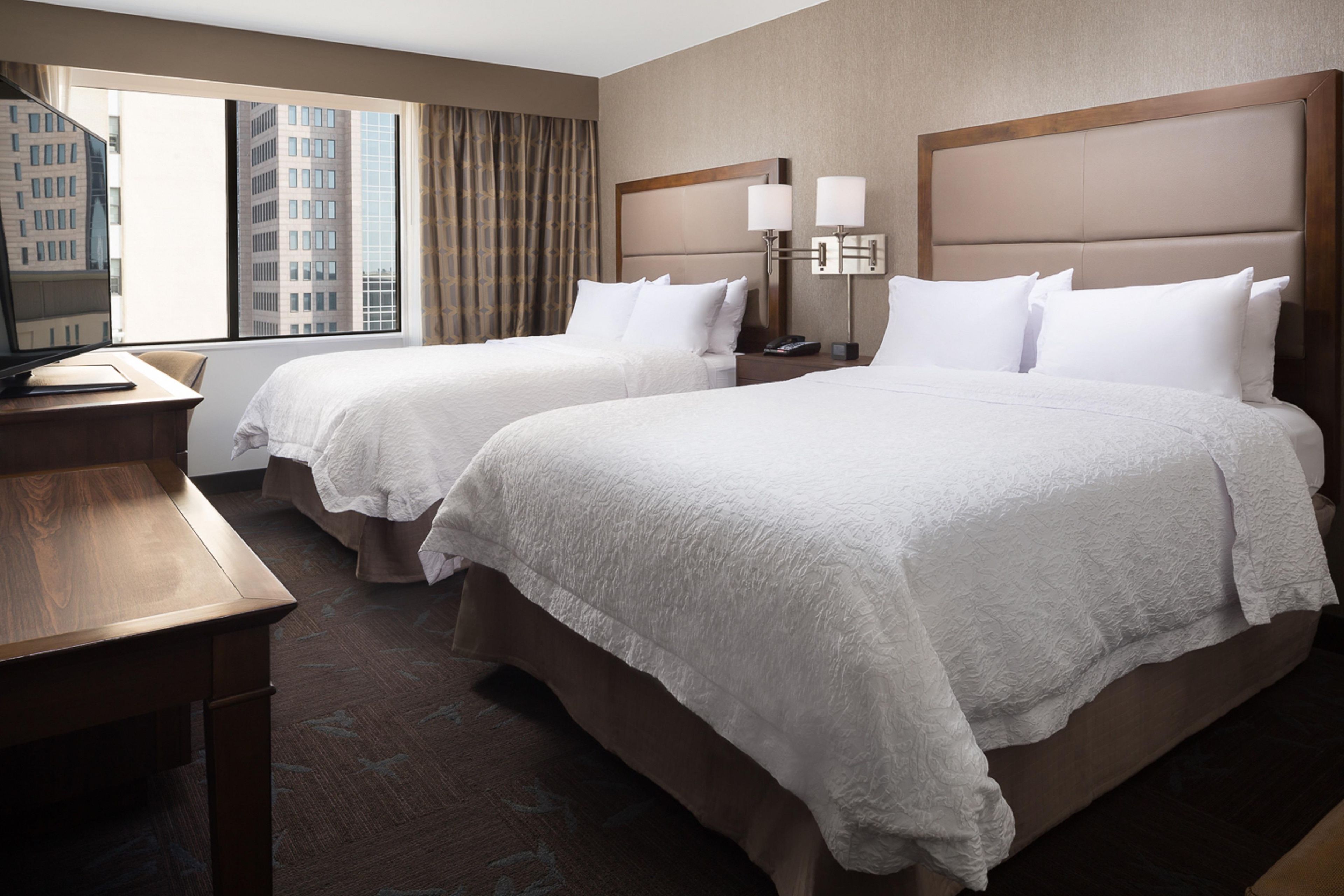A hotel room with two beds, a desk, and a window with a view of buildings.