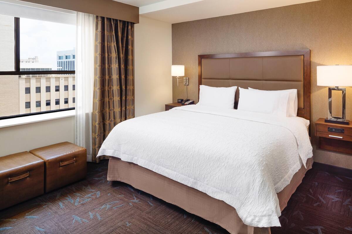 A hotel room with a large bed, a nightstand with a lamp, a small seating area, and a window with curtains.